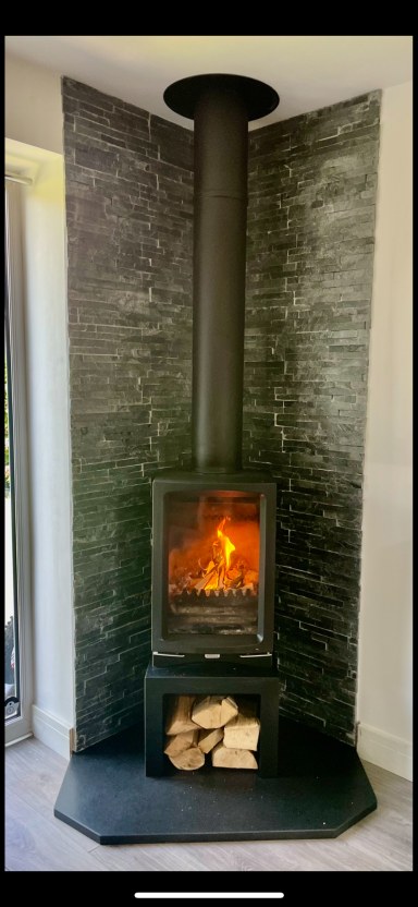 Slate-clad corner wood burner with a lit fire and log store, Bristol install